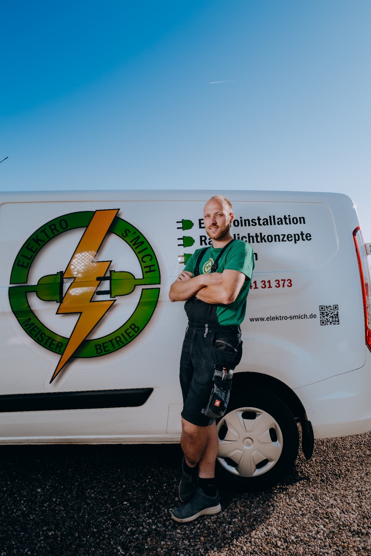 Elektriker von Elektro-s'Mich mit verschränkten Armen vor dem weißen Firmenwagen mit Logo